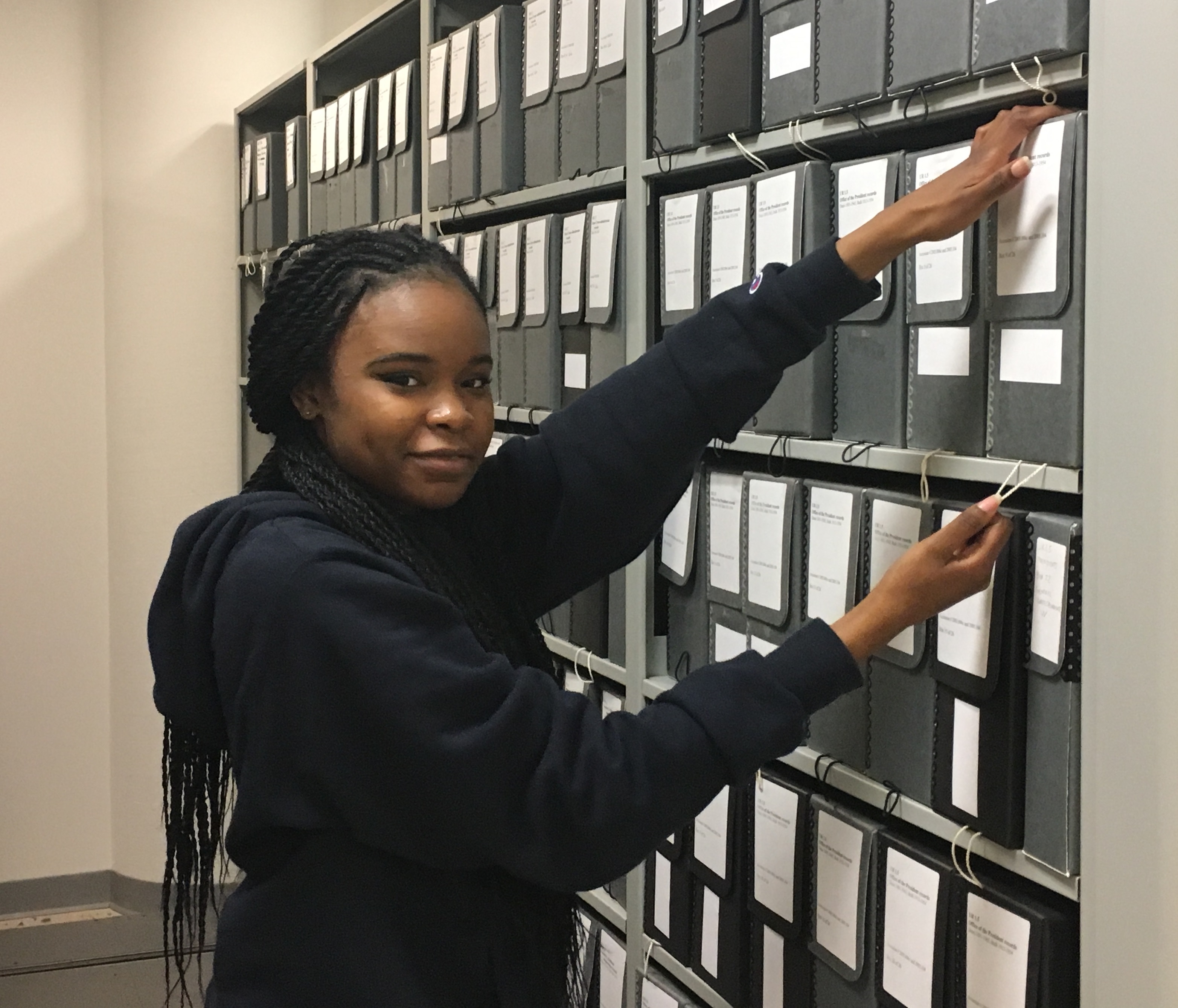 A young woman poses in front of shelves lined with folders of archival materials